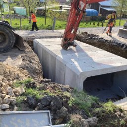 F.H.U KAM-DRÓG Kamil Mazur - Betonowy przepust skrzynkowy umieszczany w wykopie za pomocą koparki, widoczni pracownicy w odblaskowych kamizelkach i sprzęt geodezyjny w tle, teren budowy w słoneczny dzień.