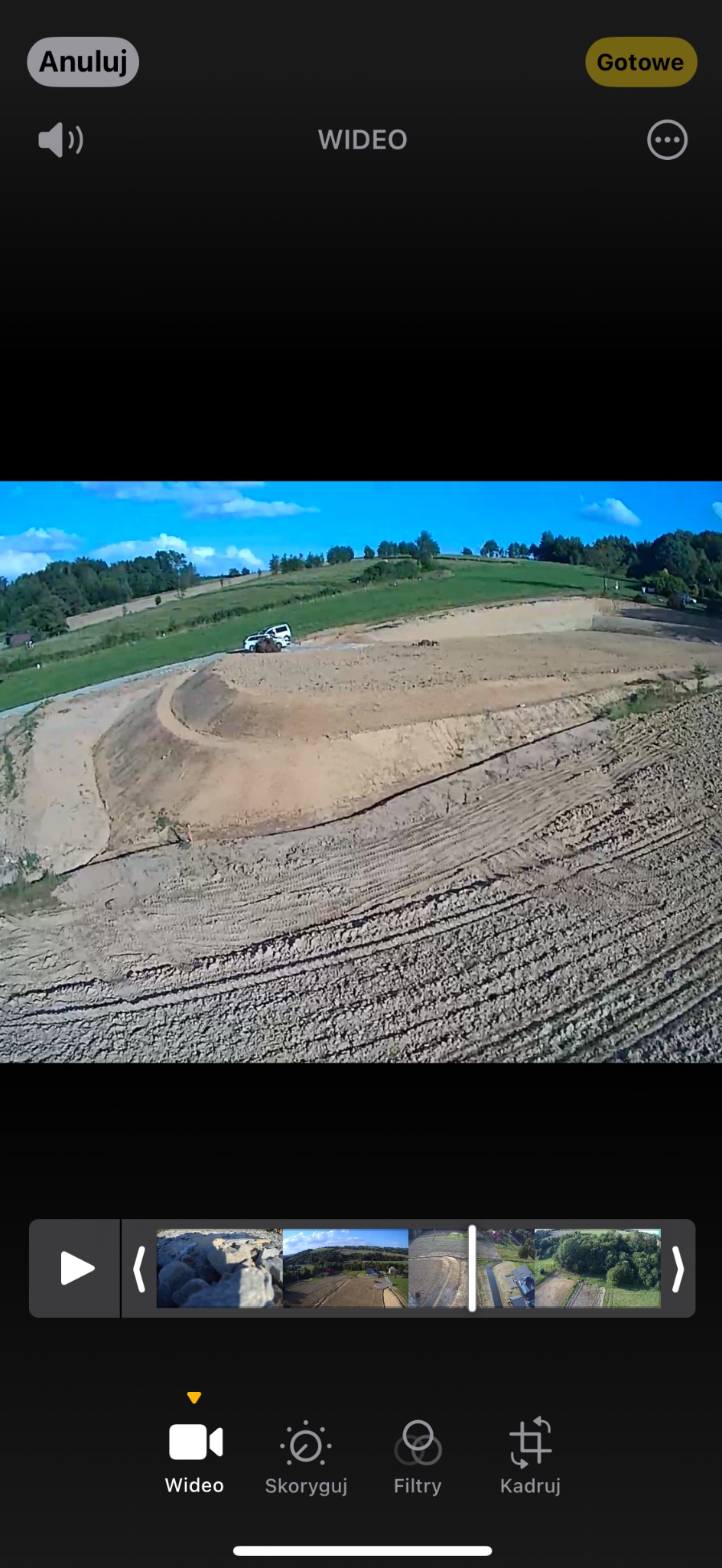 Niwelacja terenu pod budowę drogi na wzgórzu, widoczne ślady opon i dwa samochody na poboczu, w tle zielone łąki i las.