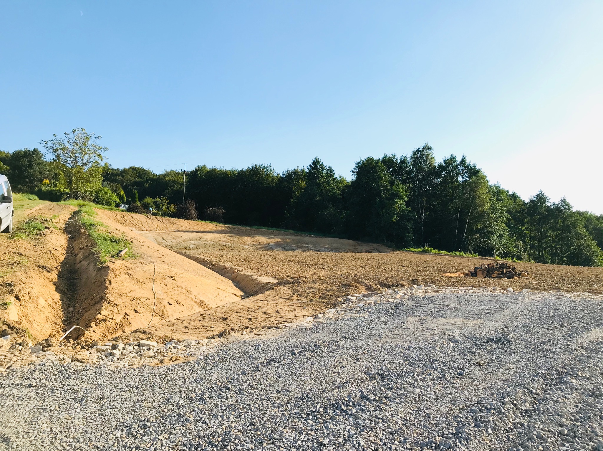 Przygotowany teren pod budowę z widocznym wykopem i utwardzoną żwirową drogą dojazdową, w tle zalesiony obszar i błękitne niebo.
