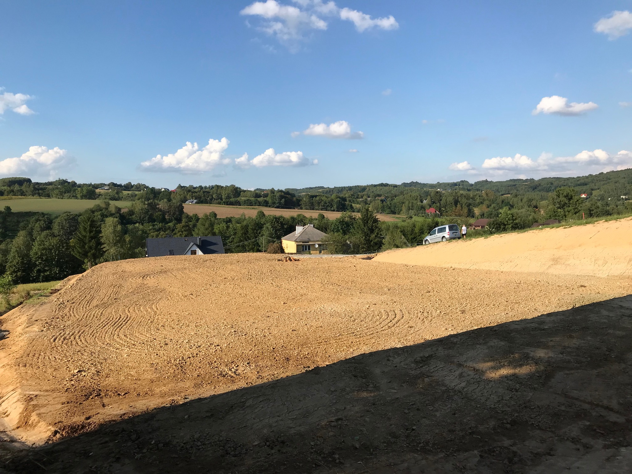 Równy, przygotowany teren pod budowę z widocznymi śladami po maszynach, w tle zabudowa wiejska i błękitne niebo z chmurami.