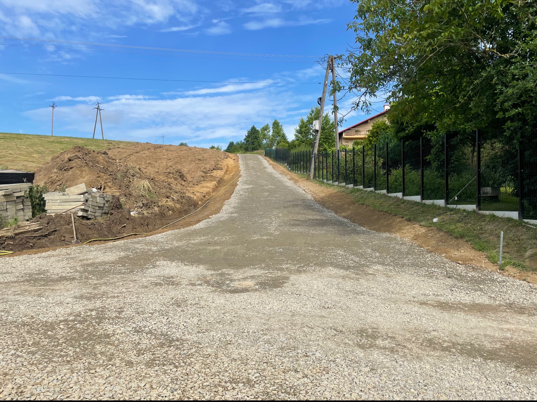 Droga gruntowa z kruszywa prowadząca pod górę, obok czarne ogrodzenie i wykopana ziemia, błękitne niebo z chmurami w tle.
