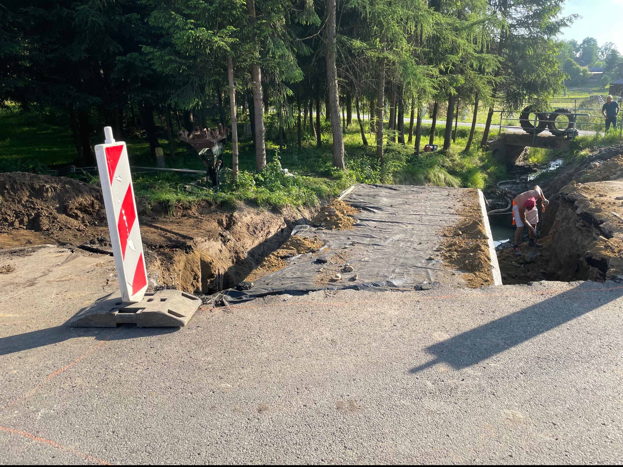 Przygotowanie podłoża pod drogę: wykopany rów zabezpieczony czarną folią, robotnicy pracujący w wykopie, w tle mostek z oponami, znak ostrzegawczy po lewej stronie.
