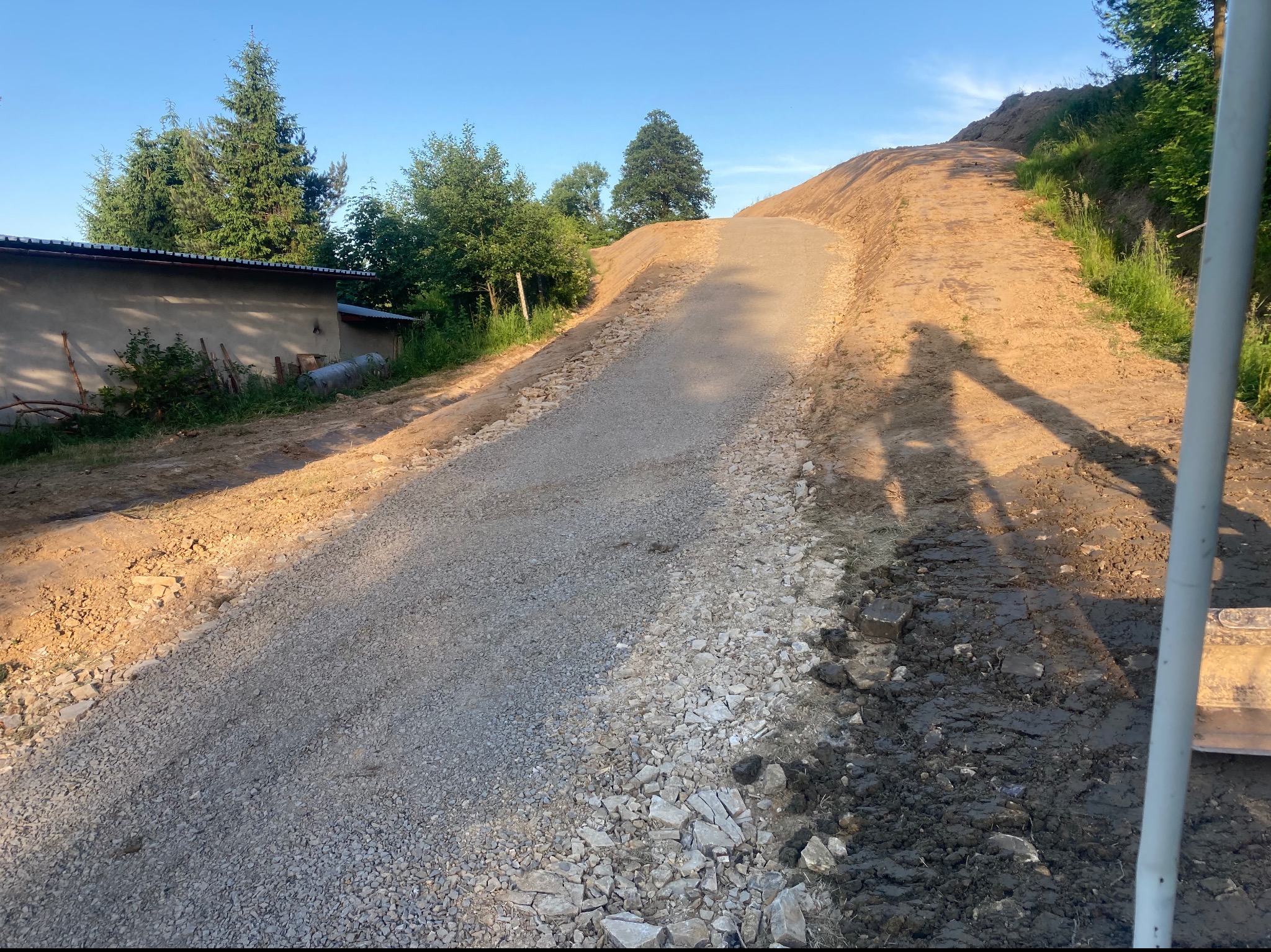 Przygotowany teren pod budowę drogi z widoczną warstwą kamienia łupanego i frakcji 4/32 kliniec, otoczony ziemią i roślinnością, w tle budynek gospodarczy i drzewa pod błękitnym niebem.