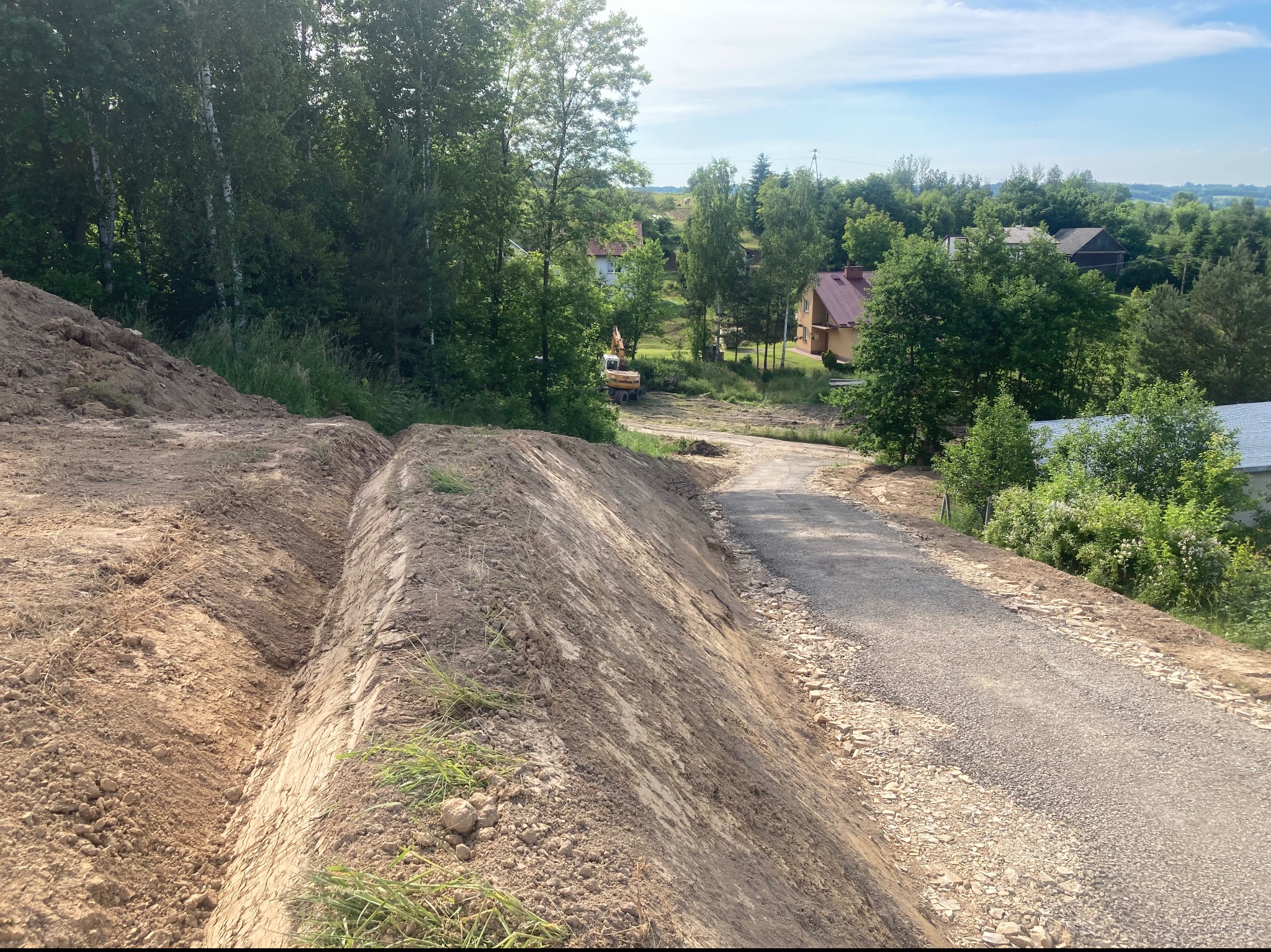 Przygotowanie terenu pod drogę: wykopane rowy po lewej stronie, w tle widoczny żółty sprzęt budowlany oraz zabudowania mieszkalne w otoczeniu zieleni.
