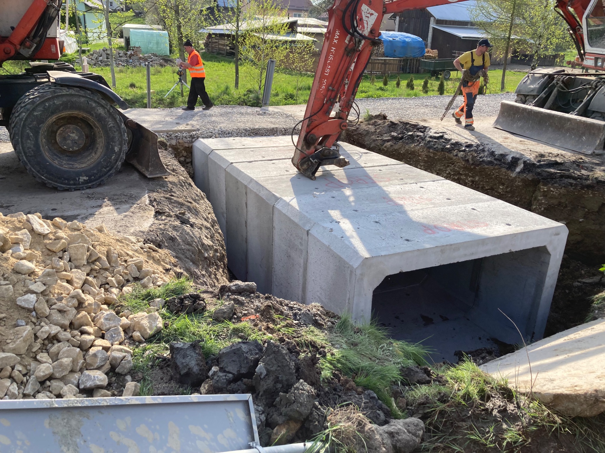 Betonowy przepust skrzynkowy umieszczany w wykopie za pomocą koparki, widoczni pracownicy w odblaskowych kamizelkach i sprzęt geodezyjny w tle, teren budowy w słoneczny dzień.