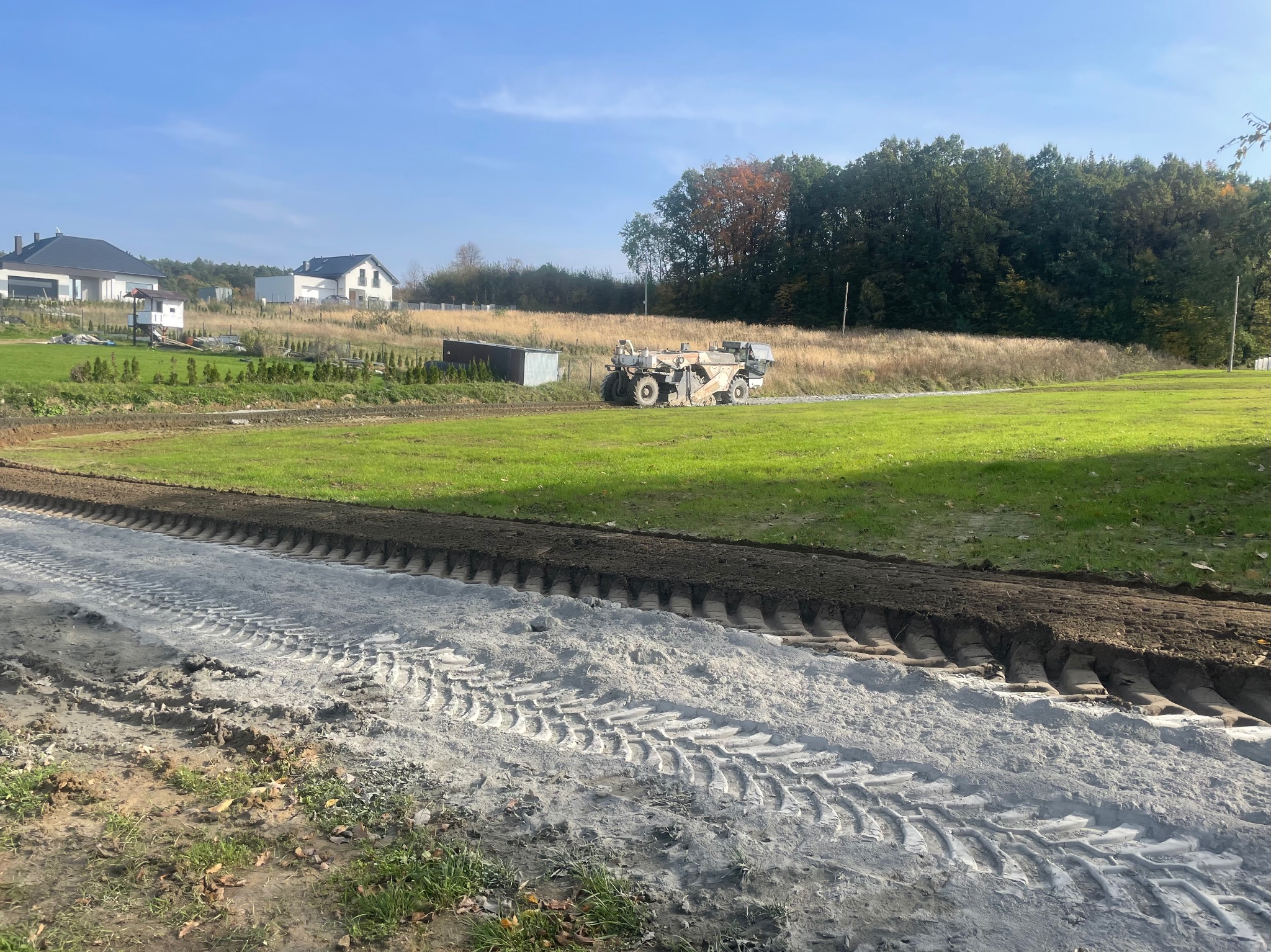 Specjalistyczna maszyna do stabilizacji gruntu cementem pracuje na placu budowy drogi, widoczne ślady opon i świeżo przygotowany teren pod nawierzchnię, w tle domy jednorodzinne i drzewa.