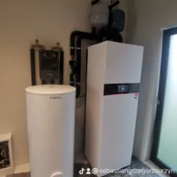 Biały zasobnik ciepłej wody i jednostka grzewcza z czarnymi rurami i zaworami, zainstalowane w pomieszczeniu z zabezpieczoną folią podłogą i widocznym oknem.