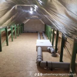 Instalacja wentylacji w surowym poddaszu: widoczne izolowane rury wentylacyjne, centrala wentylacyjna i drewniana konstrukcja dachu z folią termoizolacyjną.