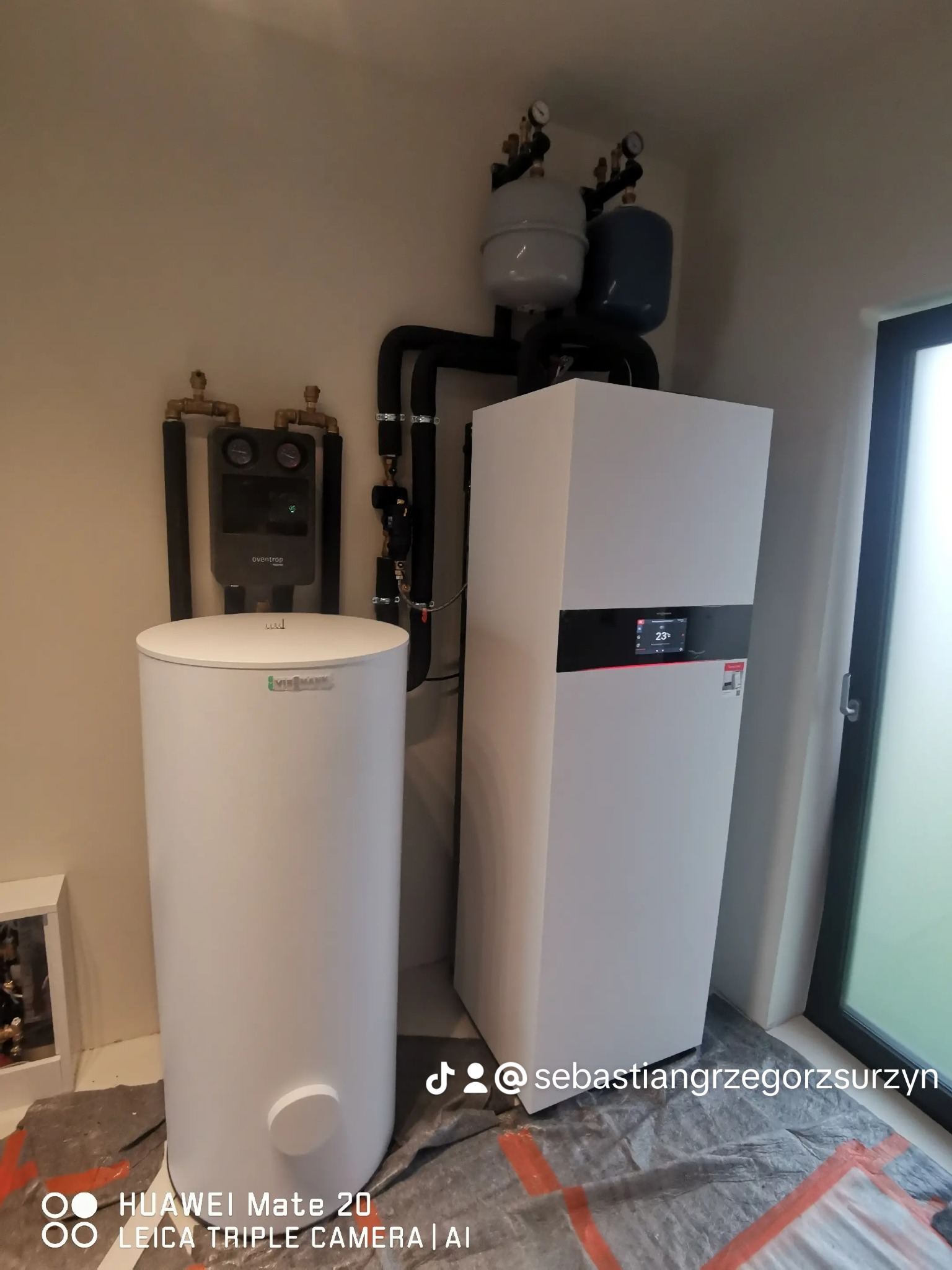 Biały zasobnik ciepłej wody i jednostka grzewcza z czarnymi rurami i zaworami, zainstalowane w pomieszczeniu z zabezpieczoną folią podłogą i widocznym oknem.