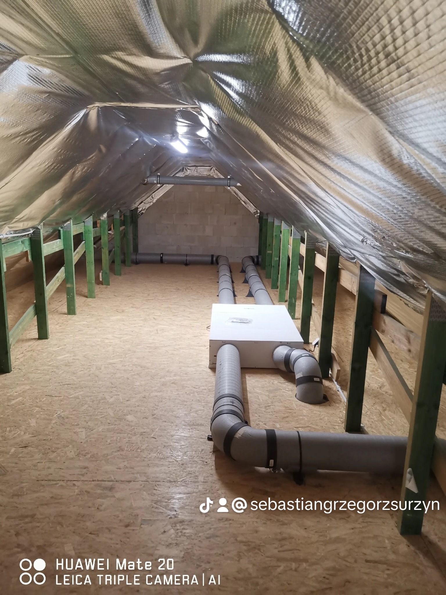 Instalacja wentylacji w surowym poddaszu: widoczne izolowane rury wentylacyjne, centrala wentylacyjna i drewniana konstrukcja dachu z folią termoizolacyjną.