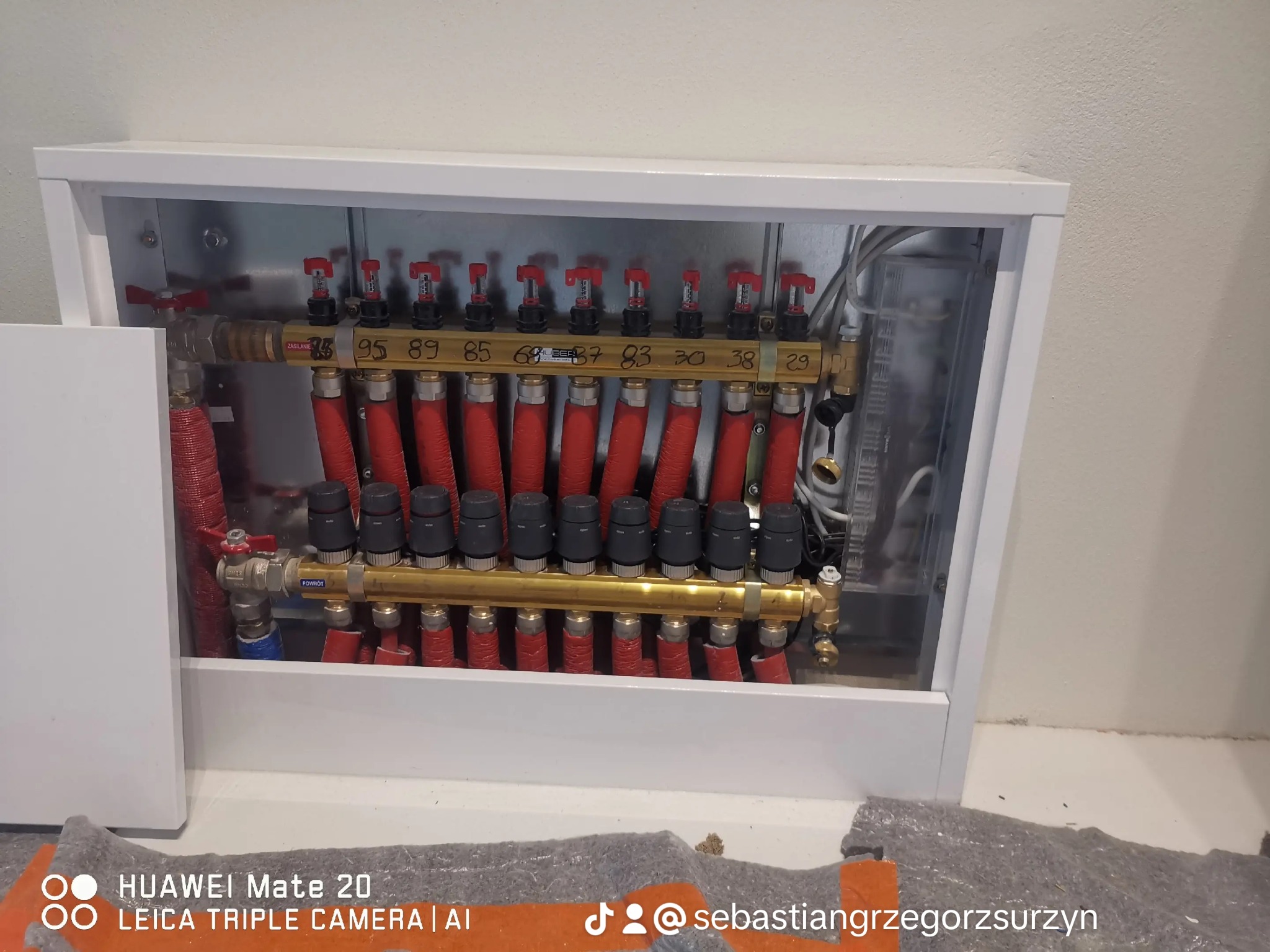 Rozdzielacz ogrzewania podłogowego w białej szafce, widoczne rury w izolacji, zawory z czerwonymi pokrętłami i czarne termostaty, numery na belce rozdzielacza.