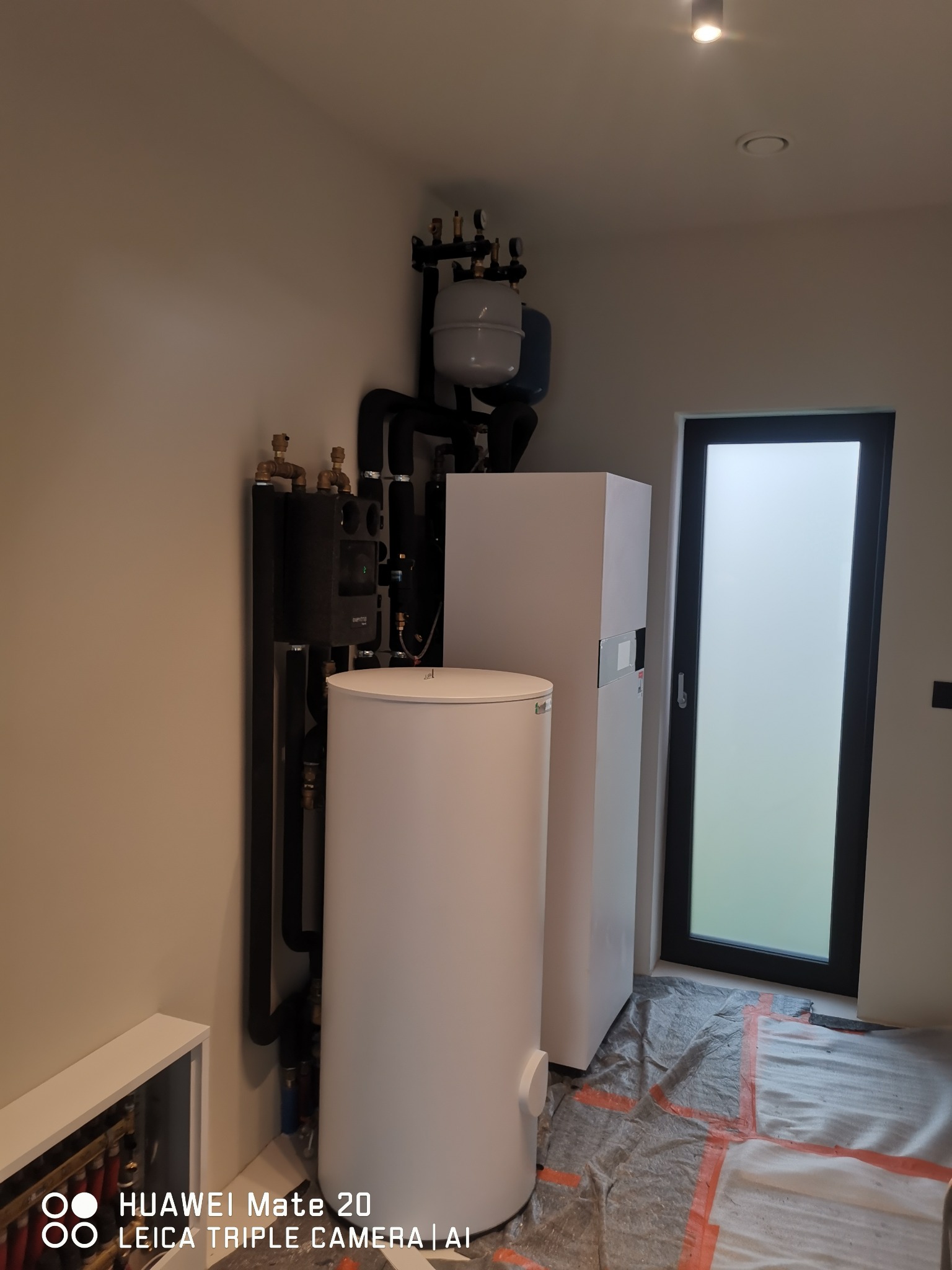 Kompaktowy zestaw grzewczy z białym zasobnikiem i czarną armaturą, zainstalowany w pomieszczeniu z oknem, podłoga zabezpieczona folią malarską, widoczny fragment rozdzielacza ogrzewania podłogowego.