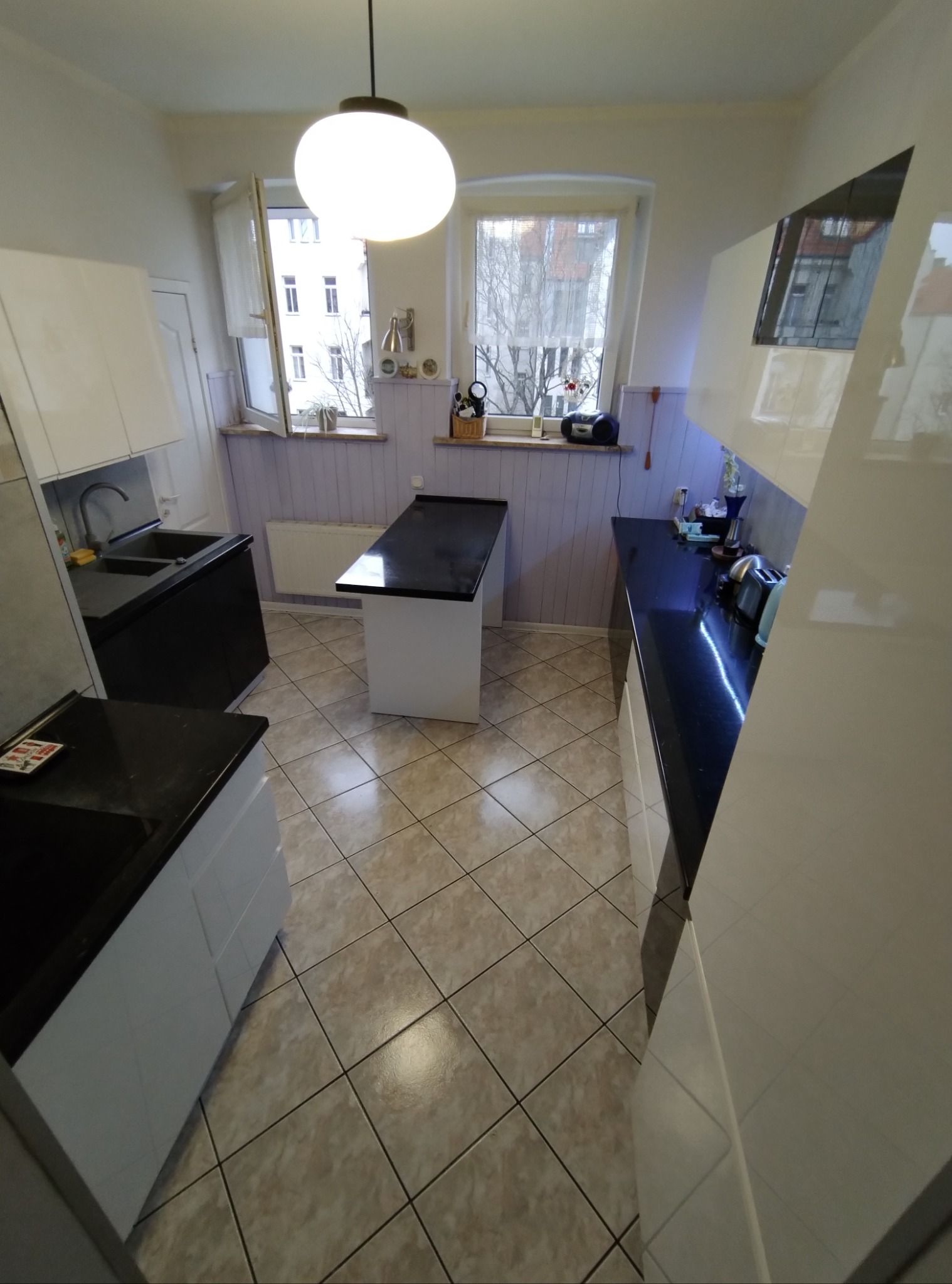 Umyta kuchnia z czarnymi blatami i białymi szafkami, widok z góry na czyste płytki podłogowe i okno z widokiem na budynek. Świeżo posprzątane wnętrze.