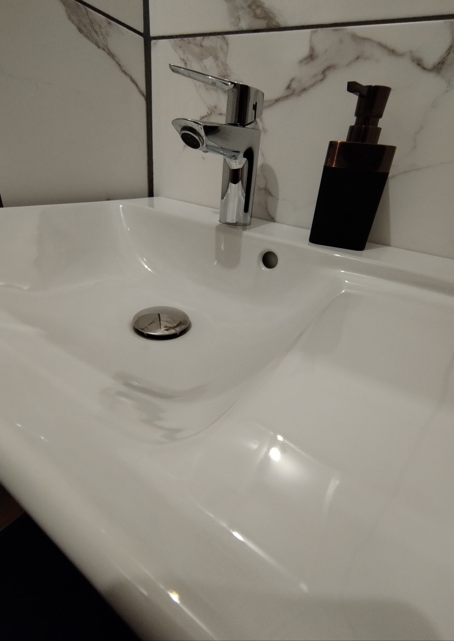 Biała, ceramiczna umywalka z chromowaną baterią i dozownikiem mydła na tle marmurowych płytek. Ujęcie z bliska, prezentujące czystość i nowoczesny design.