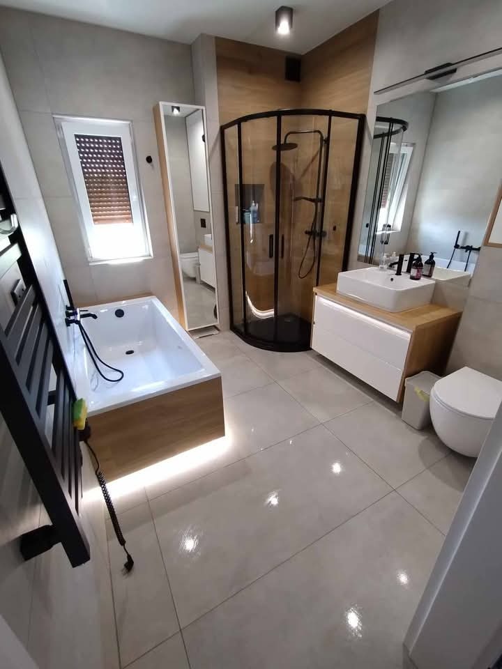 Nowoczesna łazienka z kabiną prysznicową typu walk-in, podświetlaną wanną i minimalistyczną umywalką. Jasne płytki i drewniane akcenty tworzą elegancki wystrój.
