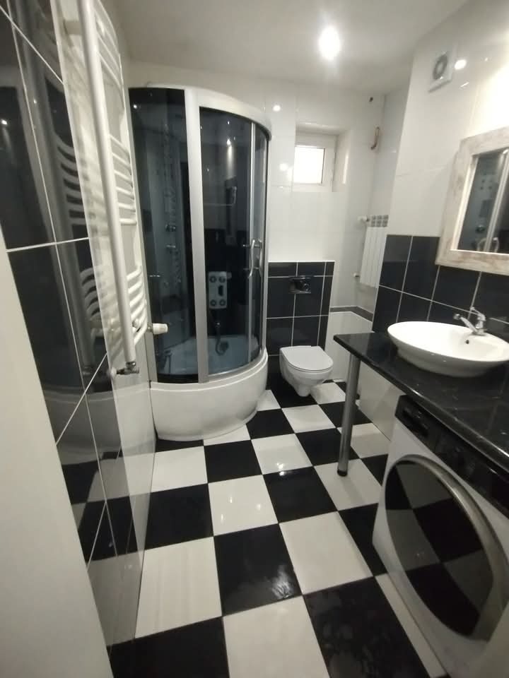 Błyszcząca, czysto biała kabina prysznicowa z chromowanymi detalami w odświeżonej łazience z biało-czarną szachownicą na podłodze i ciemnym blatem.