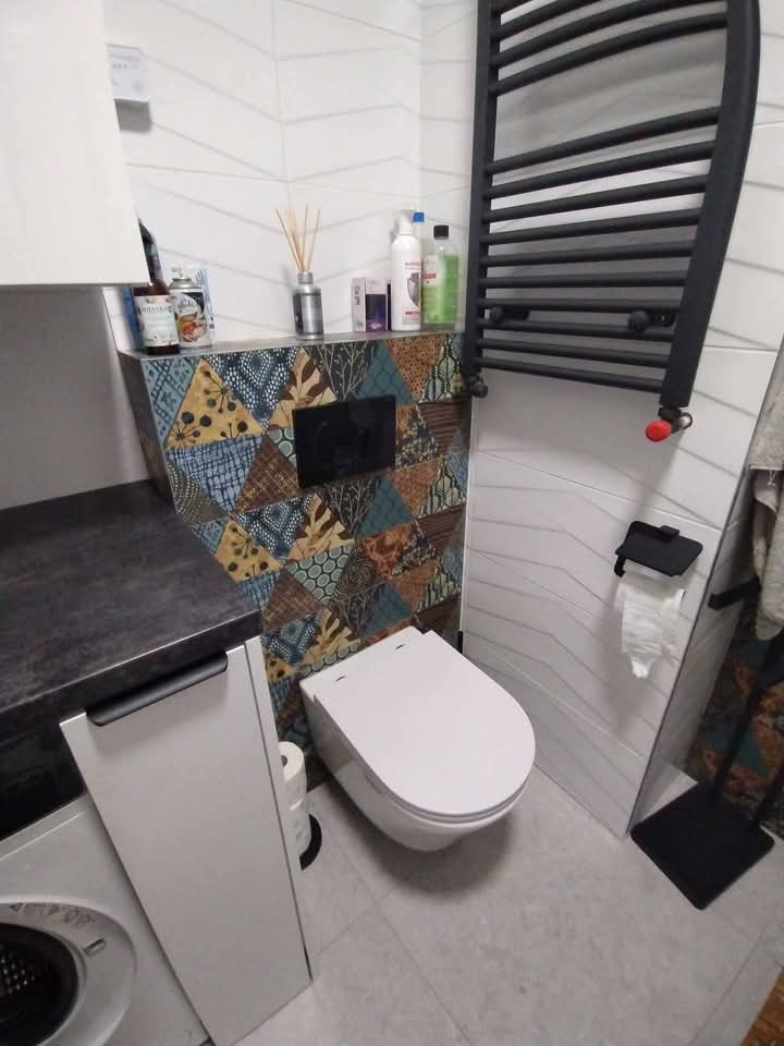 Umywalka z szafką i toaleta w nowoczesnej łazience z wzorzystą ścianą i grzejnikiem drabinkowym. Widoczne akcesoria łazienkowe i pralka.