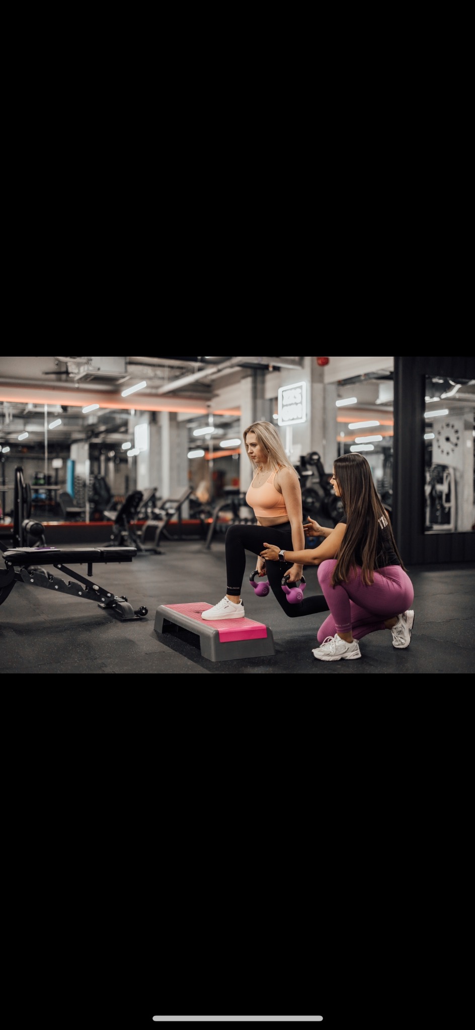 Kobieta wykonuje wypad w przód z obciążeniem na stepie, pod nadzorem drugiej kobiety w siłowni.