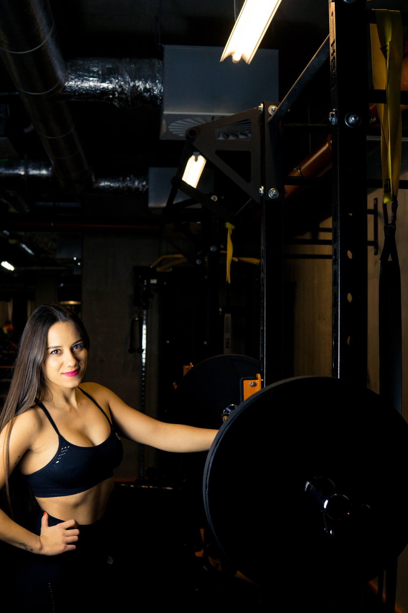 Kobieta w sportowym stroju pozuje obok sztangi na tle wyposażenia siłowni, z widocznymi rurami wentylacyjnymi na suficie.