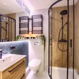 Kam-bud - Nowoczesna łazienka z kabiną prysznicową z czarnymi ramami, drewnopodobnymi płytkami na ścianach, umywalką z szafką z drewnianym fornirem, podświetlanym lustrem i grzejnikiem drabinkowym w czarnym...