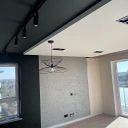 Kam-bud - Wnętrze mieszkania w trakcie wykańczania: szpachlowana ściana z wyprowadzonymi instalacjami elektrycznymi, częściowo pomalowany sufit w kolorze czarnym z reflektorami, widok na balkon z dużym oknem.