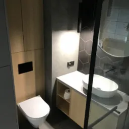 Nowoczesna łazienka z umywalką nablatową, toaletą wiszącą i kabiną prysznicową z czarnymi profilami, wykończona płytkami heksagonalnymi i drewnopodobnymi panelami.