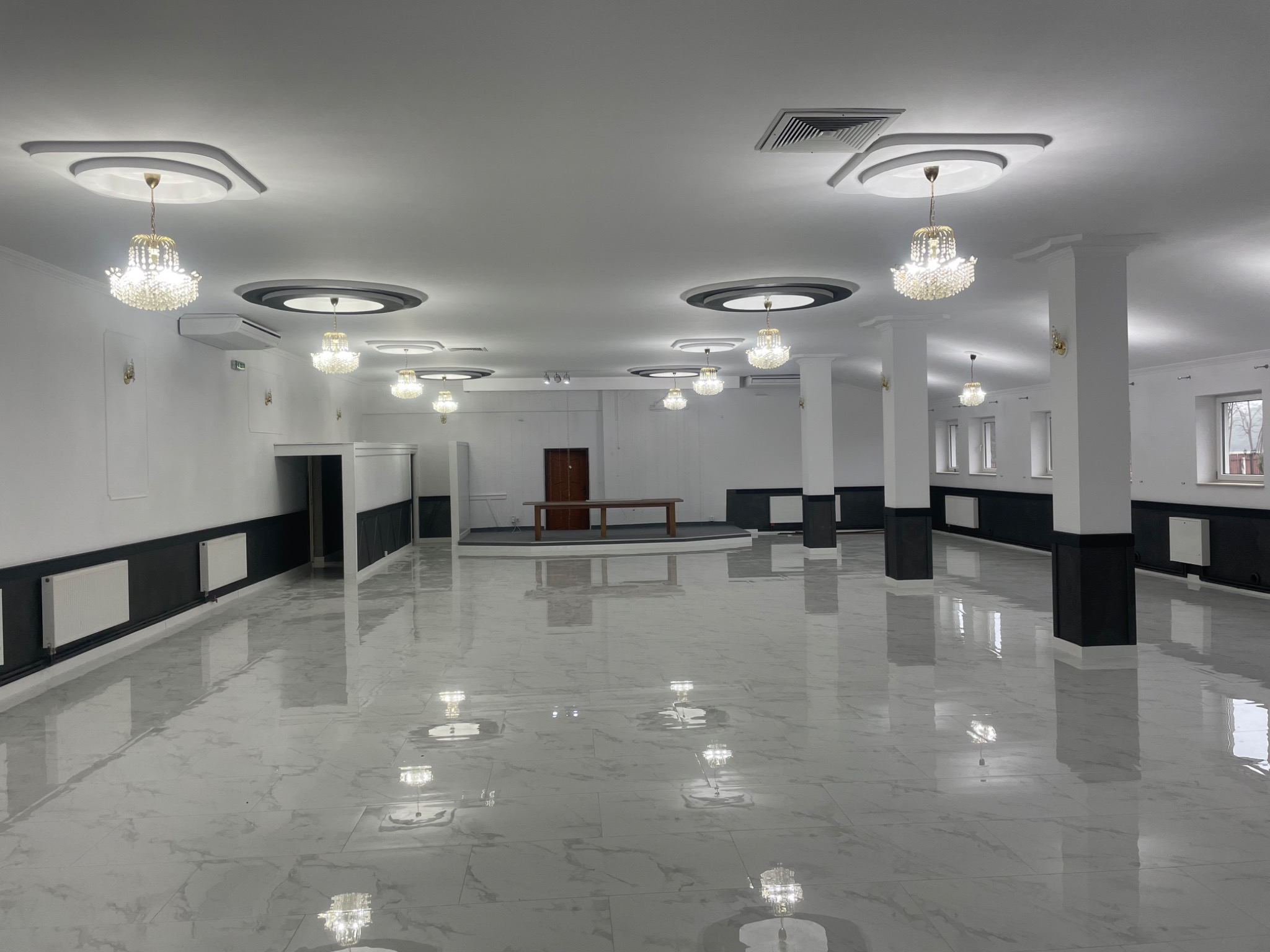 Przestronne, odnowione wnętrze sali balowej z marmurową podłogą, białymi ścianami z czarnymi akcentami i kryształowymi żyrandolami pod sufitem.