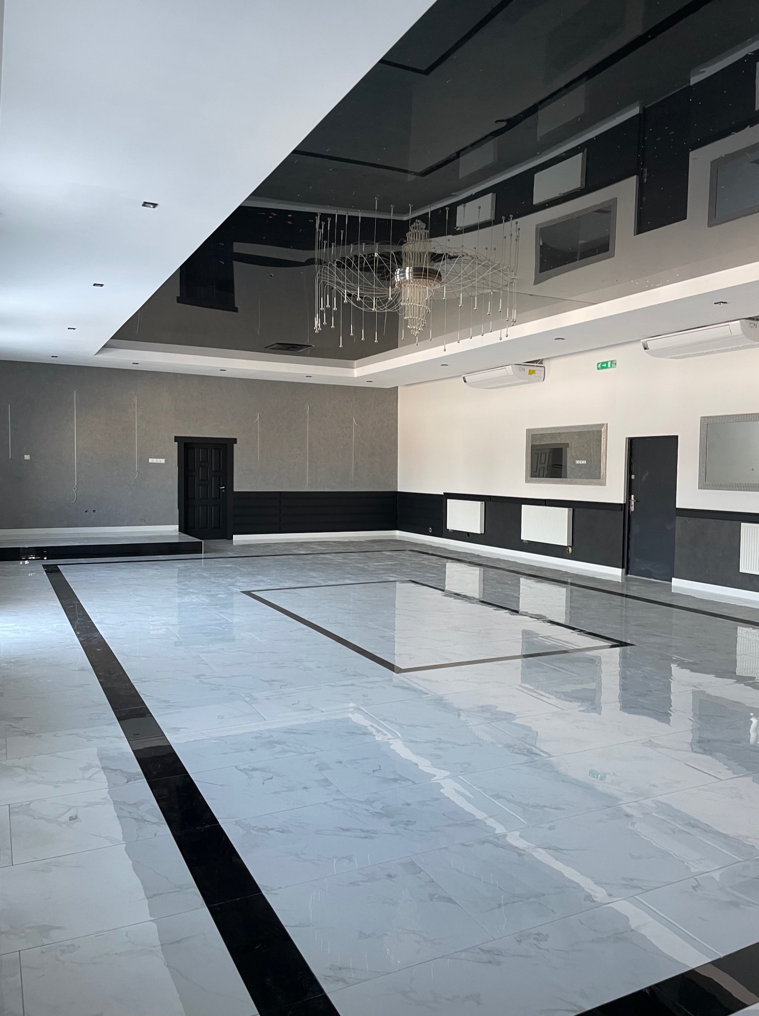 Przestronne, nowocześnie wykończone wnętrze sali z marmurową posadzką w odcieniach szarości i bieli, czarnym akcentem w postaci obramowania podłogi i sufitu, oraz dekoracyjnym żyrandolem.