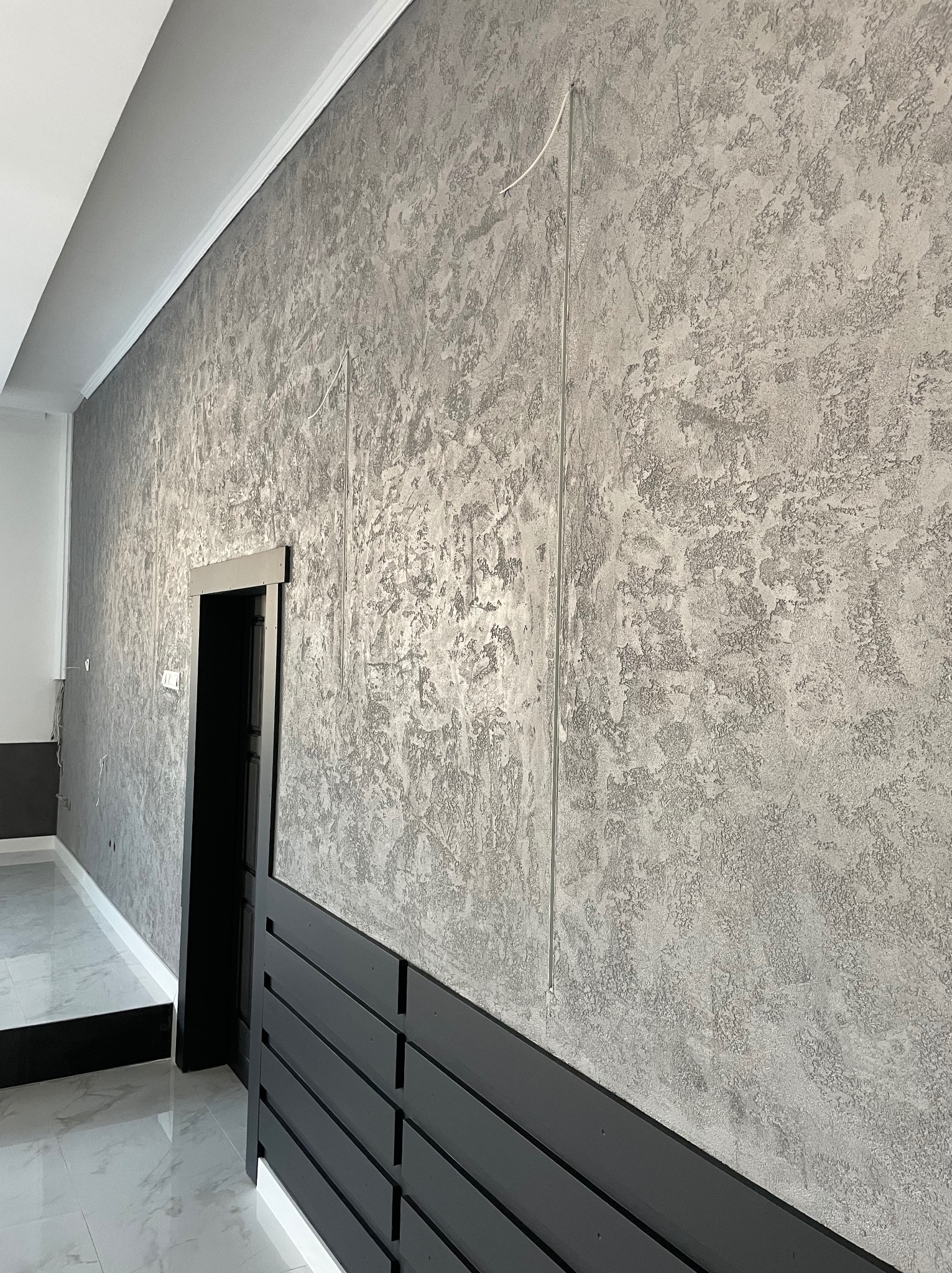 Ściana z dekoracyjną szarą fakturą, czarna listwa przypodłogowa z poziomymi szczelinami i czarne obramowanie drzwi, widoczne białe listwy sufitowe i marmurowe płytki podłogowe.