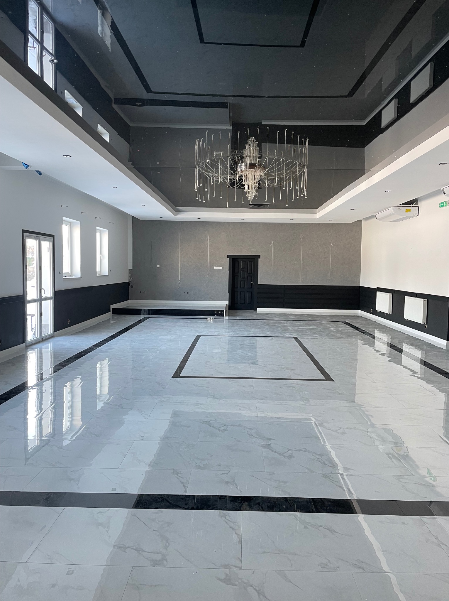 Przestronne, nowocześnie wykończone wnętrze sali z marmurową podłogą w odcieniach szarości i czerni, błyszczącym, czarnym sufitem z designerskim żyrandolem i szarą ścianą akcentującą. Widoczne...