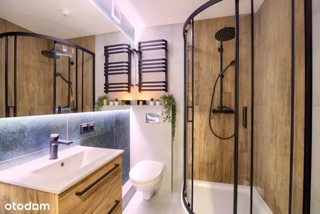 Nowoczesna łazienka z kabiną prysznicową z czarnymi ramami, drewnopodobnymi płytkami na ścianach, umywalką z szafką z drewnianym fornirem, podświetlanym lustrem i grzejnikiem drabinkowym w czarnym...