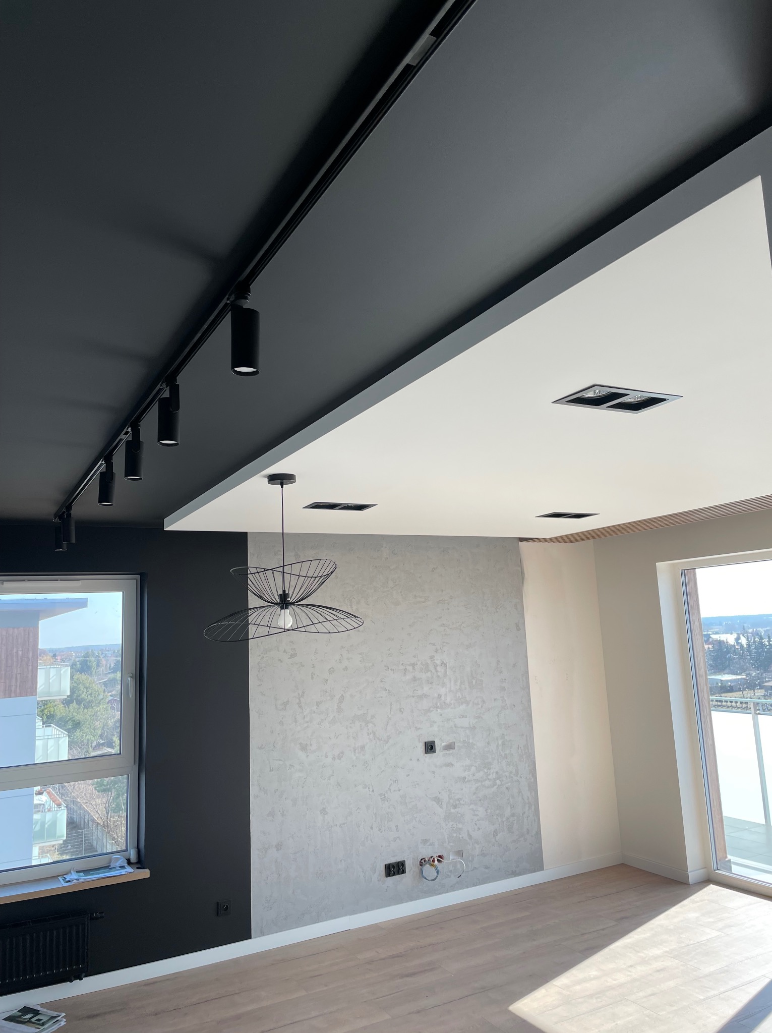 Wnętrze po remoncie: czarny sufit z reflektorami szynowymi, biały sufit z wpuszczanymi lampami, ściana z dekoracyjnym szpachlowaniem, oraz wisząca lampa druciana. Widoczne gniazdka elektryczne...