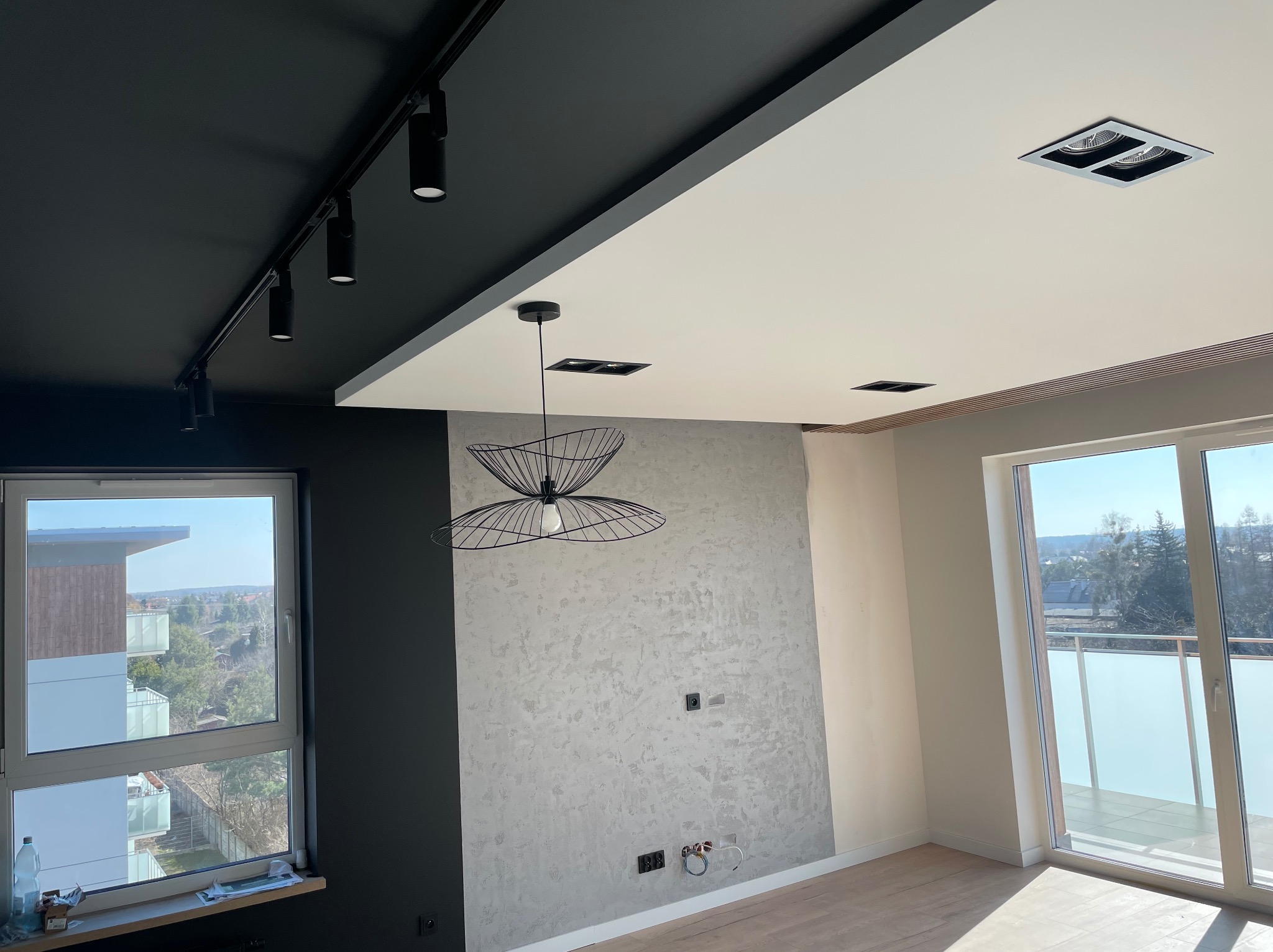 Wnętrze mieszkania w trakcie wykańczania: szpachlowana ściana z wyprowadzonymi instalacjami elektrycznymi, częściowo pomalowany sufit w kolorze czarnym z reflektorami, widok na balkon z dużym oknem.