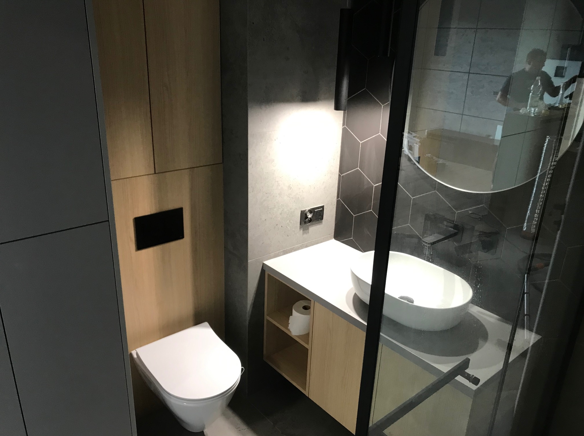 Nowoczesna łazienka z umywalką nablatową, toaletą wiszącą i kabiną prysznicową z czarnymi profilami, wykończona płytkami heksagonalnymi i drewnopodobnymi panelami.