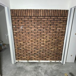 Krzysztof Bednarczyk - Ściana wykończona cegłą dekoracyjną w trakcie montażu, widoczne kable elektryczne wystające ze ściany, narzędzia w skrzynce na podłodze, surowy beton na podłodze.