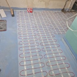 Krzysztof Bednarczyk - Montaż ogrzewania podłogowego Warmtec w Radomiu: czerwone kable grzewcze ułożone na siatce, przygotowane do zalania wylewką, widoczna puszka instalacyjna Klimas i narzędzia.