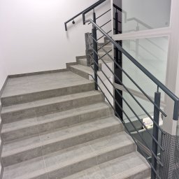 Krzysztof Bednarczyk - Schody wyłożone szarymi płytkami z metalową balustradą i szybem windy w tle.