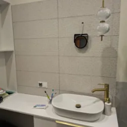 Nowoczesna łazienka z umywalką nablatową, złotą baterią i lustrem na tle ściany wyłożonej szarymi płytkami z delikatną fakturą, wisząca lampa z dwoma szklanymi kloszami.