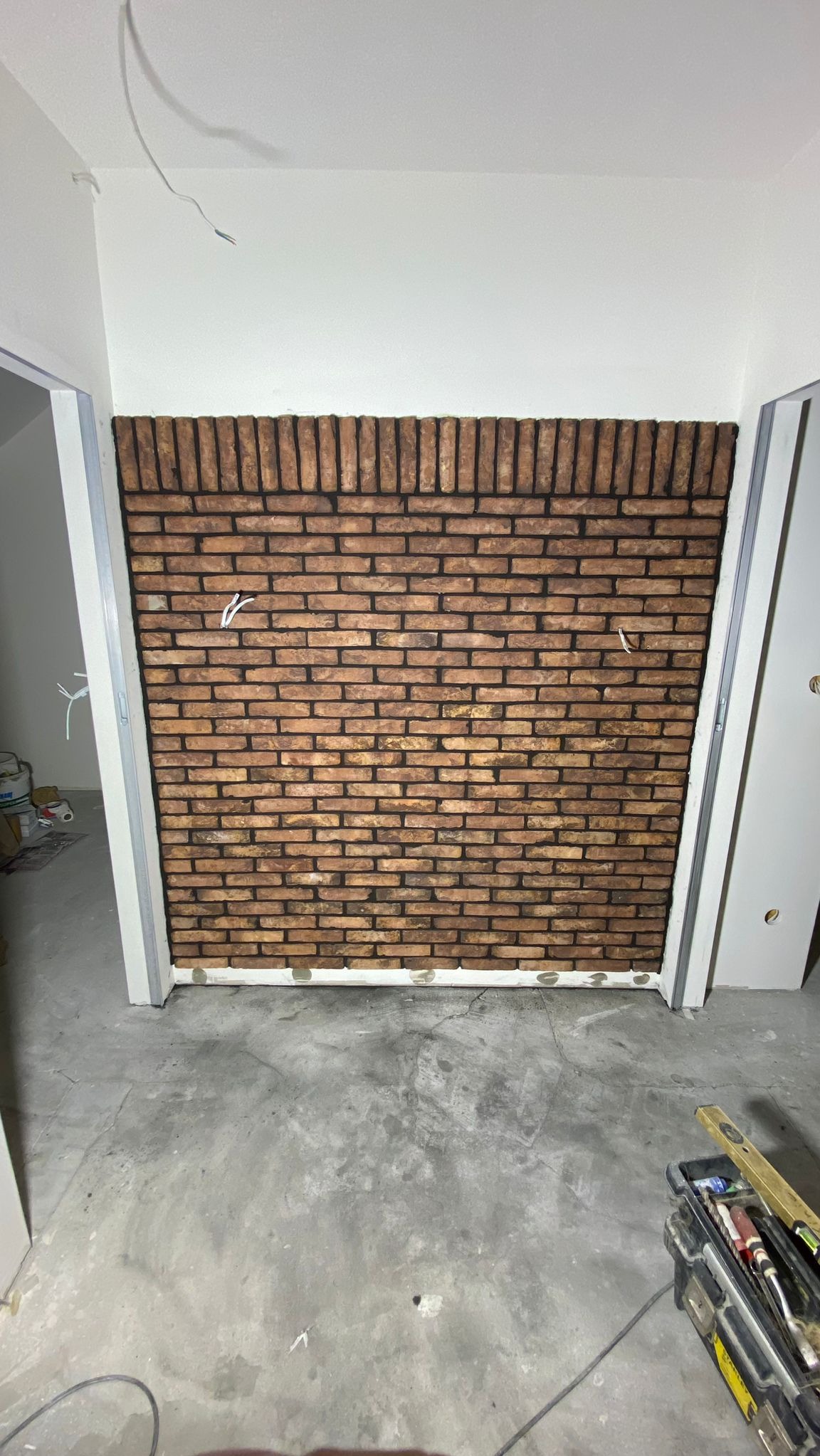 Ściana wykończona cegłą dekoracyjną w trakcie montażu, widoczne kable elektryczne wystające ze ściany, narzędzia w skrzynce na podłodze, surowy beton na podłodze.