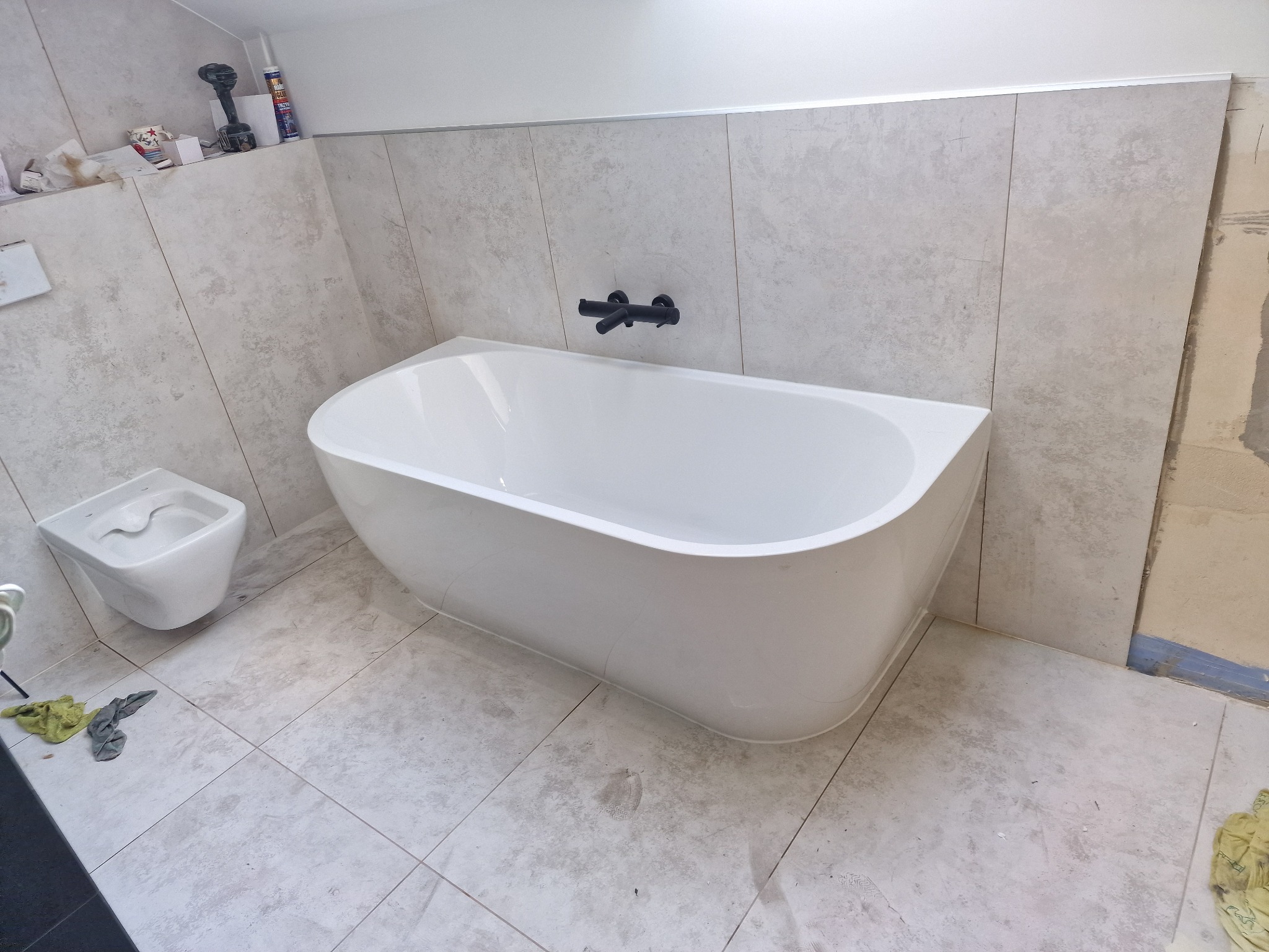Biała, wolnostojąca wanna i podwieszana toaleta w trakcie montażu w łazience wyłożonej szarymi płytkami, widoczne narzędzia i materiały budowlane.