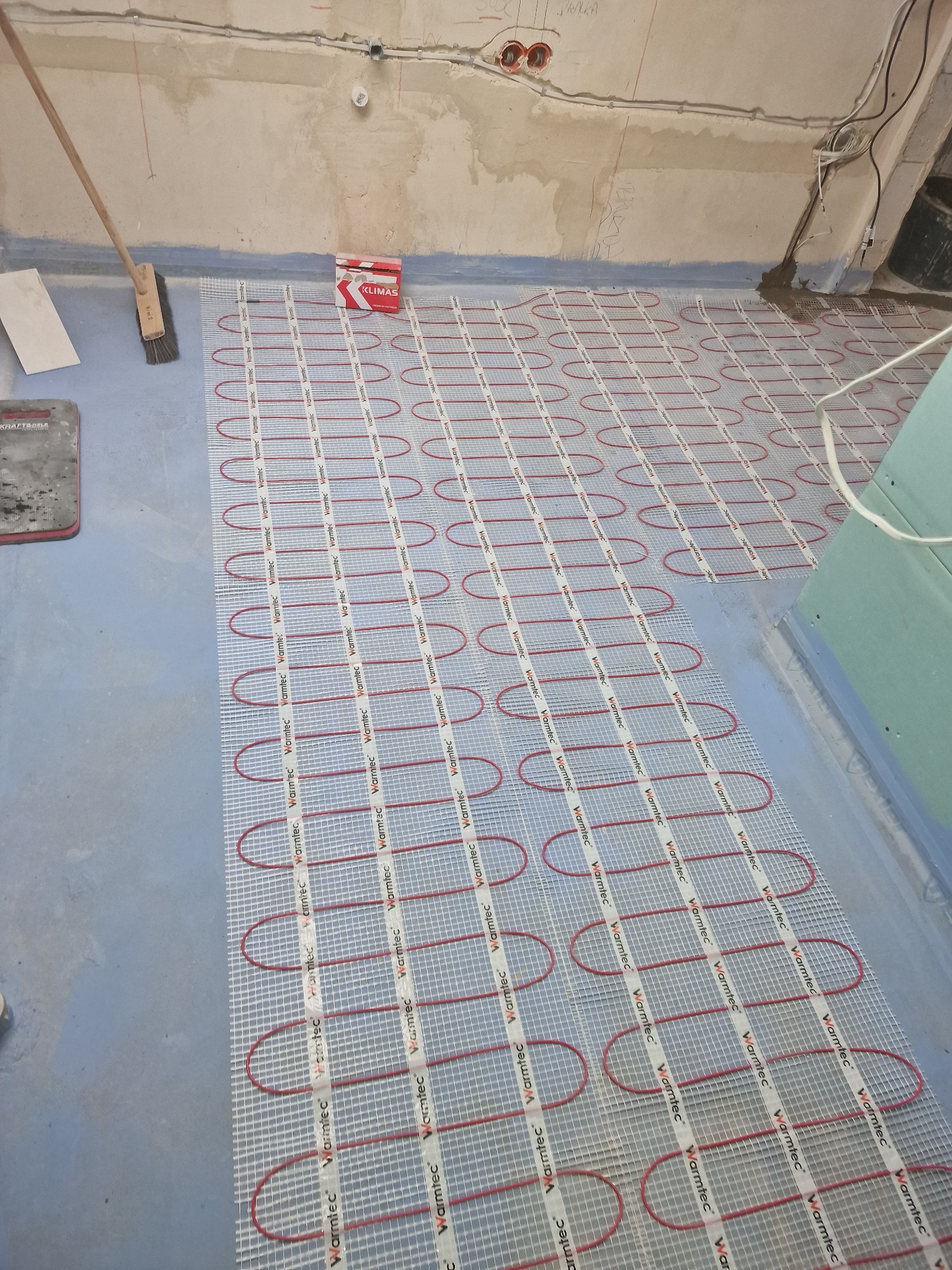 Montaż ogrzewania podłogowego Warmtec w Radomiu: czerwone kable grzewcze ułożone na siatce, przygotowane do zalania wylewką, widoczna puszka instalacyjna Klimas i narzędzia.