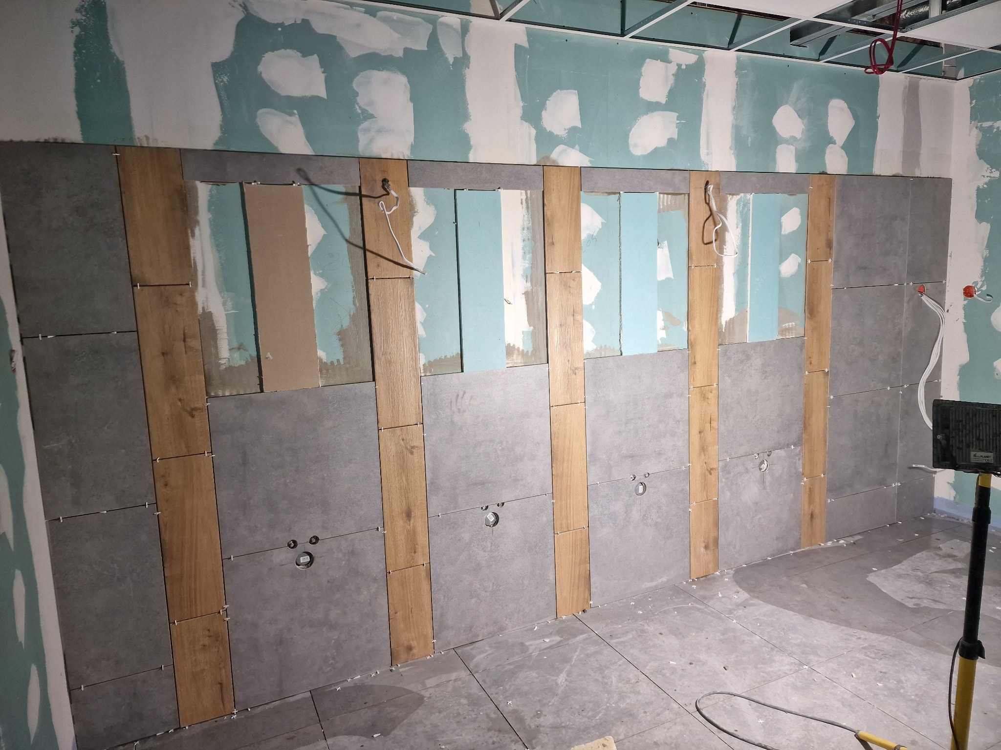 Ściana łazienki w trakcie układania płytek: szare płytki kwadratowe z fugą krzyżykową, pionowe pasy płytek imitujących drewno, widoczne otwory na instalacje, ściany przygotowane pod gładź, sufit...