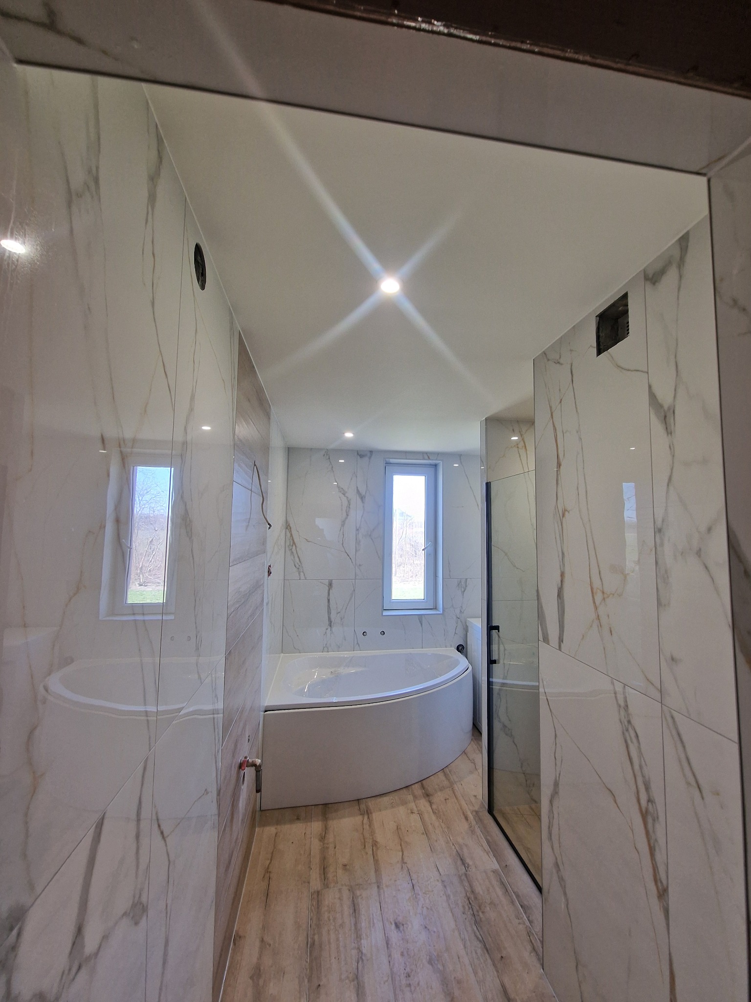 Nowoczesna łazienka z okrągłą wanną, wykończona białymi płytkami imitującymi marmur i drewnopodobną podłogą, widok z korytarza.