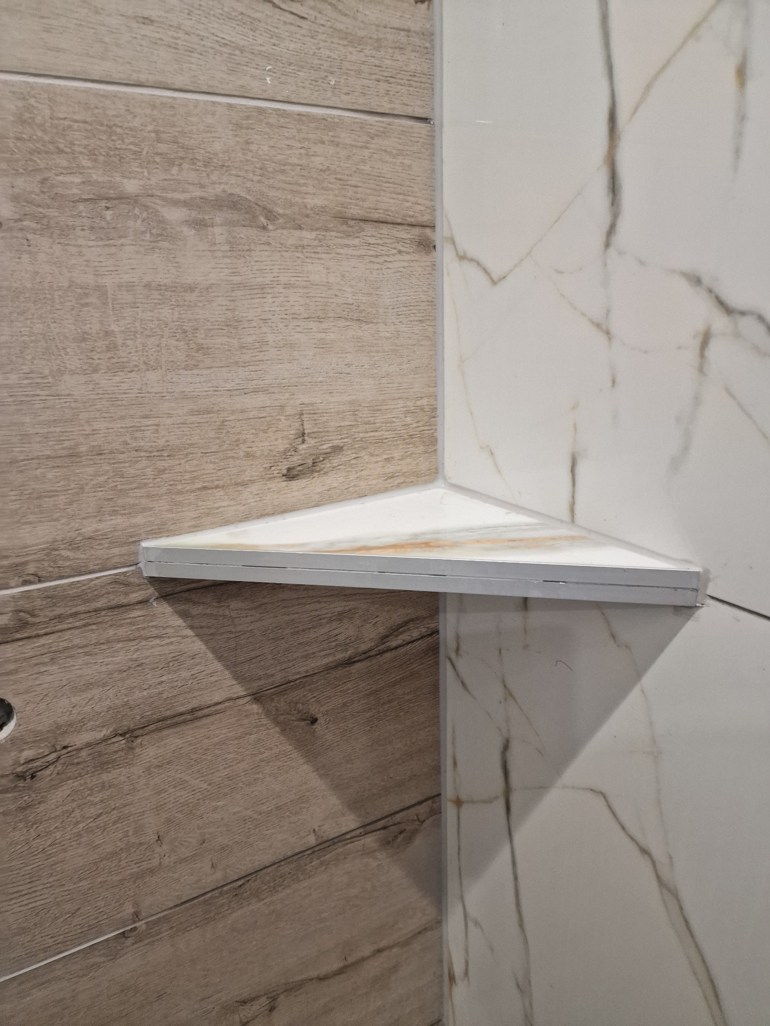 Narożna półka trójkątna z płytek ceramicznych imitujących marmur, zamontowana między ścianami z płytek drewnopodobnych i białych płytek z szarymi żyłami w łazience.