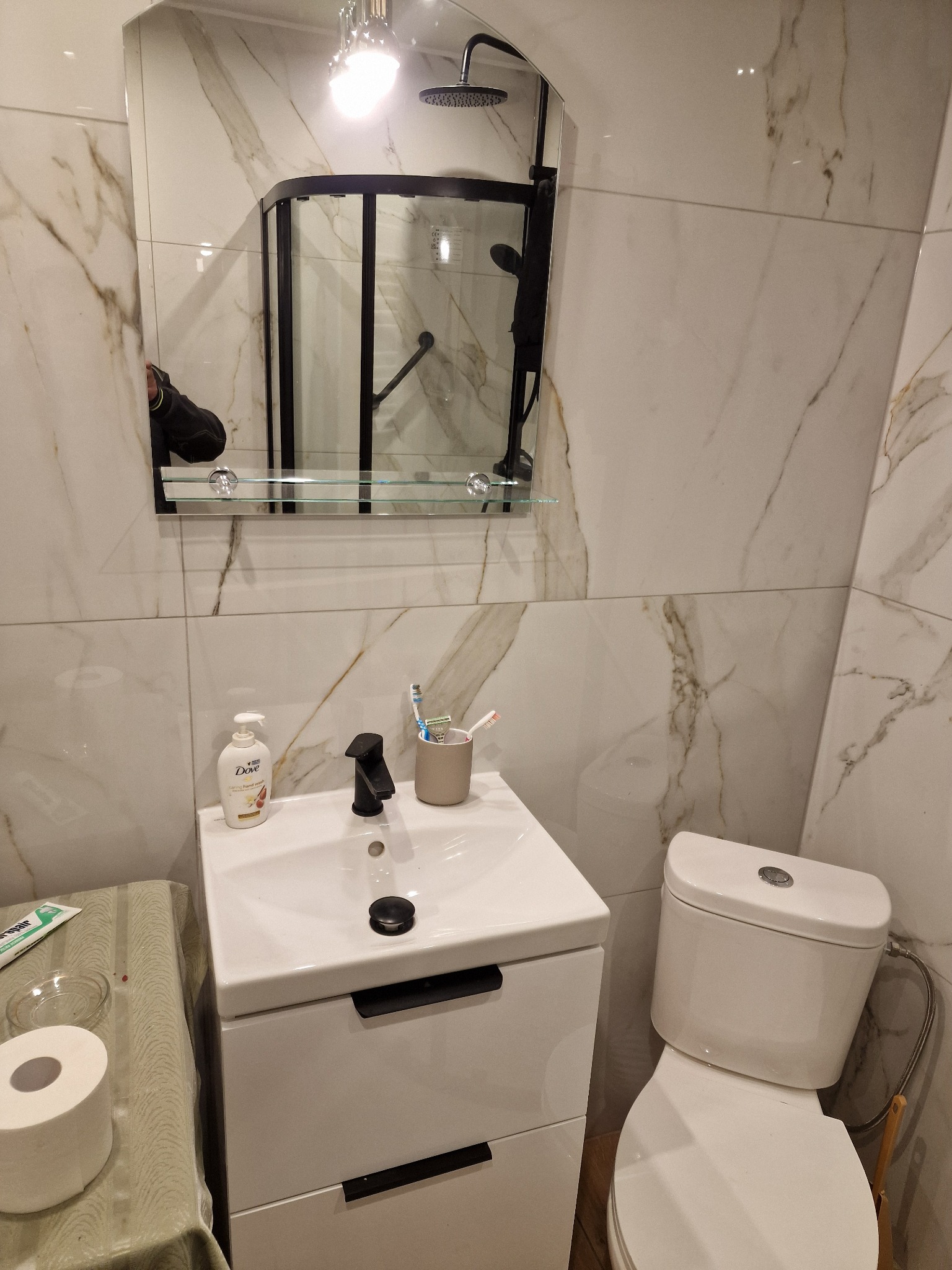 Wykończona łazienka z marmurowymi płytkami, czarną armaturą i kabiną prysznicową z czarnymi ramami, odbicie osoby w lustrze.