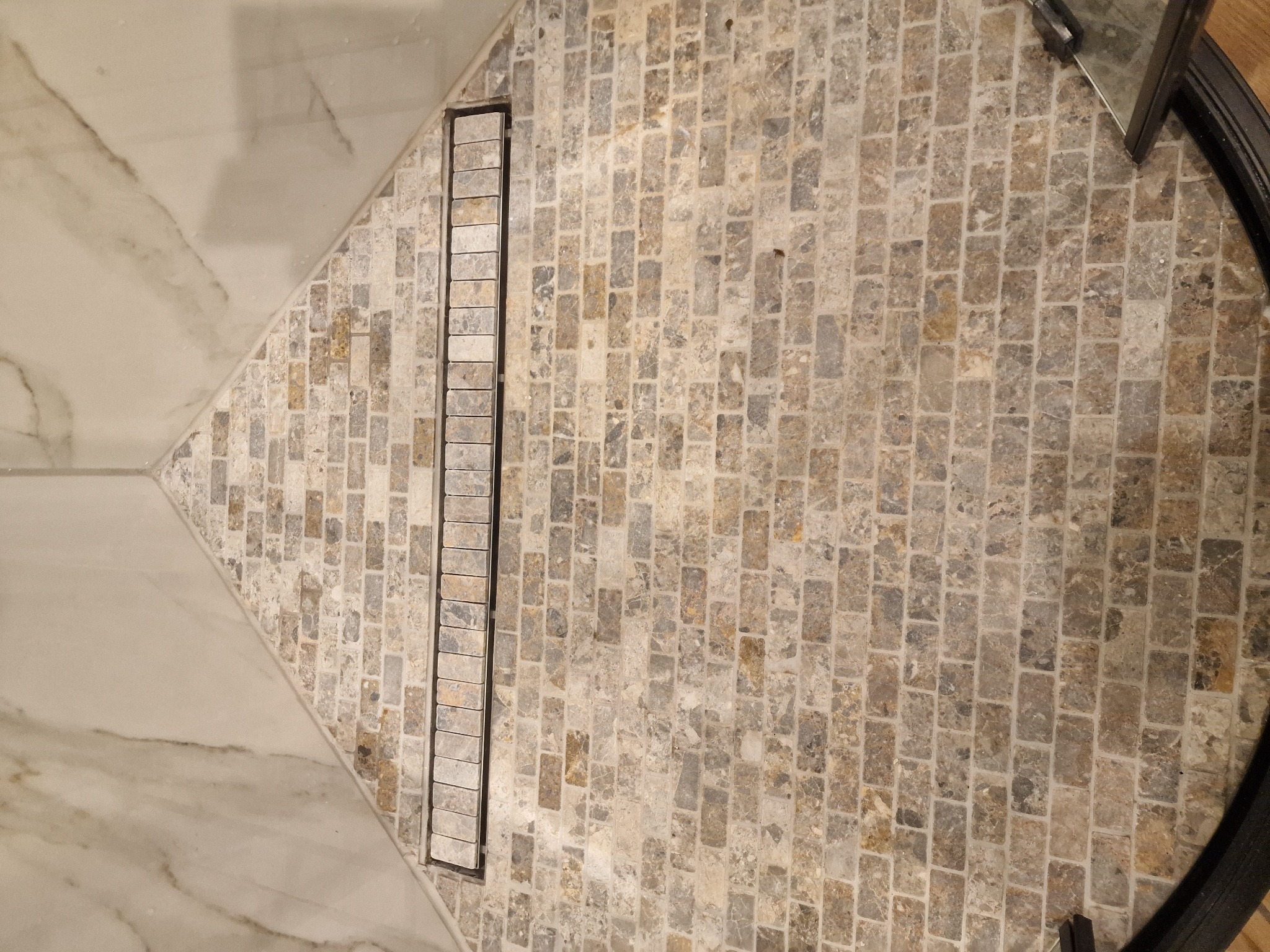 Ukończona podłoga w łazience w Radomiu, z mozaiką z kamienia naturalnego w odcieniach beżu i szarości, przylegająca do płytek imitujących marmur oraz z liniowym odpływem prysznicowym z kamienną...