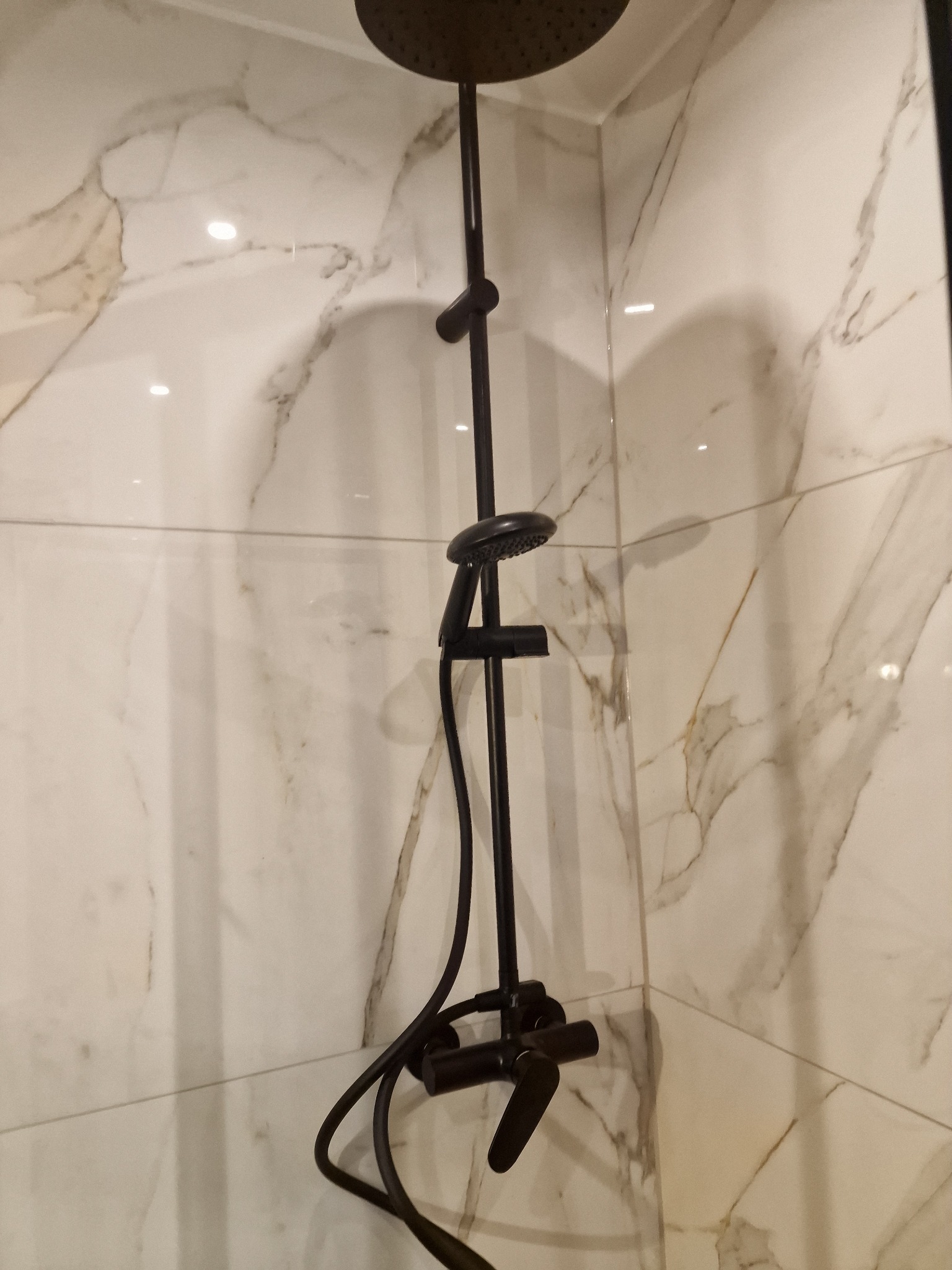 Czarny zestaw natryskowy z deszczownicą i baterią, zamontowany na ścianie wyłożonej białymi płytkami imitującymi marmur.