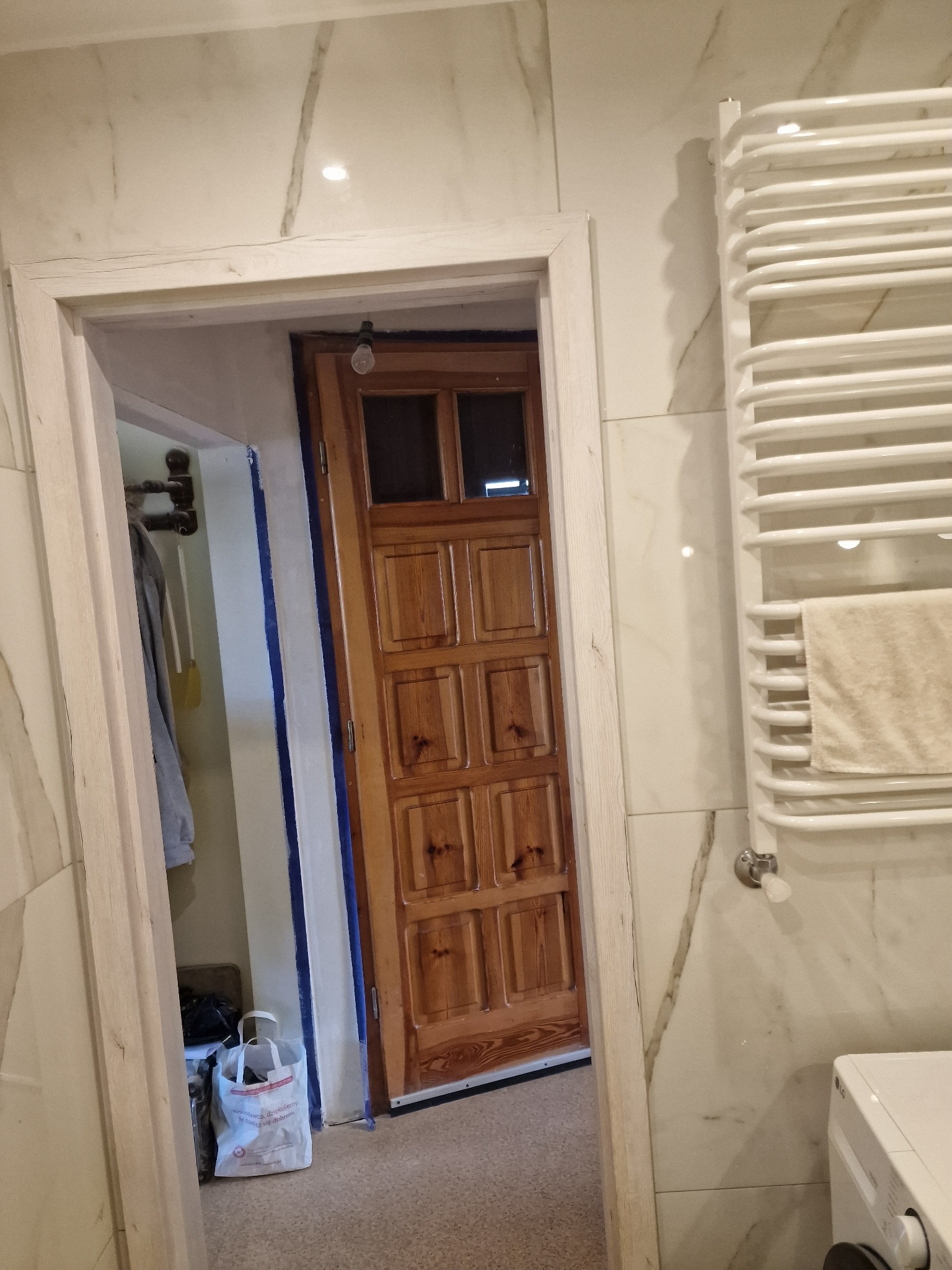 Fragment łazienki w trakcie remontu: widoczne marmurowe płytki, grzejnik drabinkowy z ręcznikiem i drewniane drzwi, zabezpieczone niebieską taśmą malarską przy futrynie.