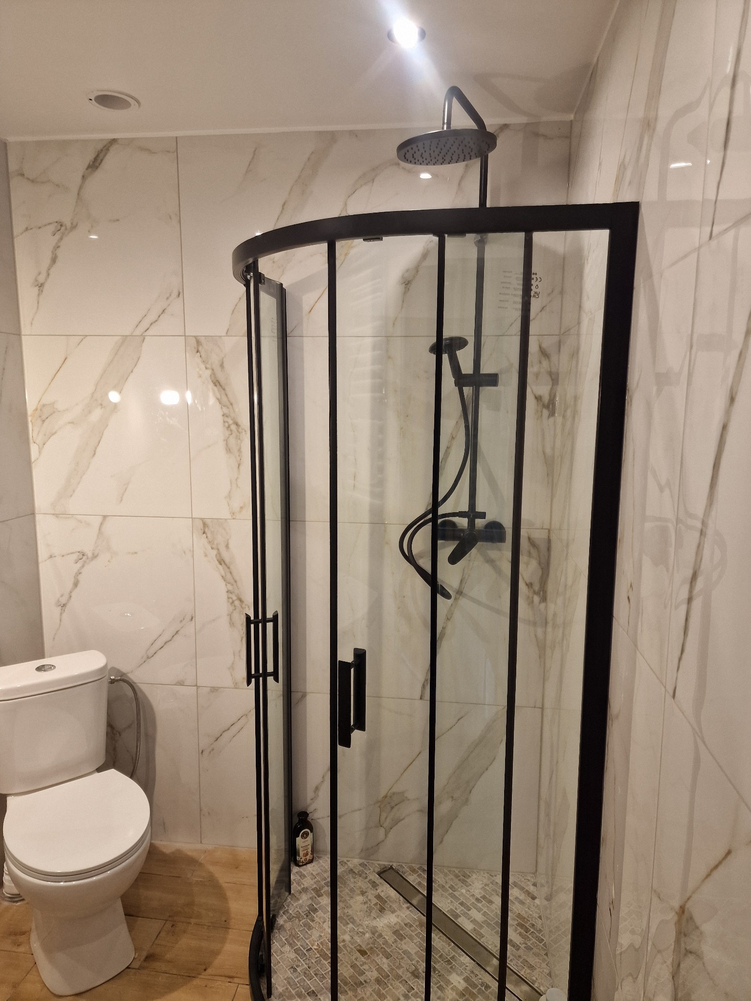 Nowoczesna kabina prysznicowa z czarną ramą i deszczownicą, jasne płytki imitujące marmur na ścianach, widoczna toaleta po lewej stronie. Drobna mozaika na podłodze kabiny.