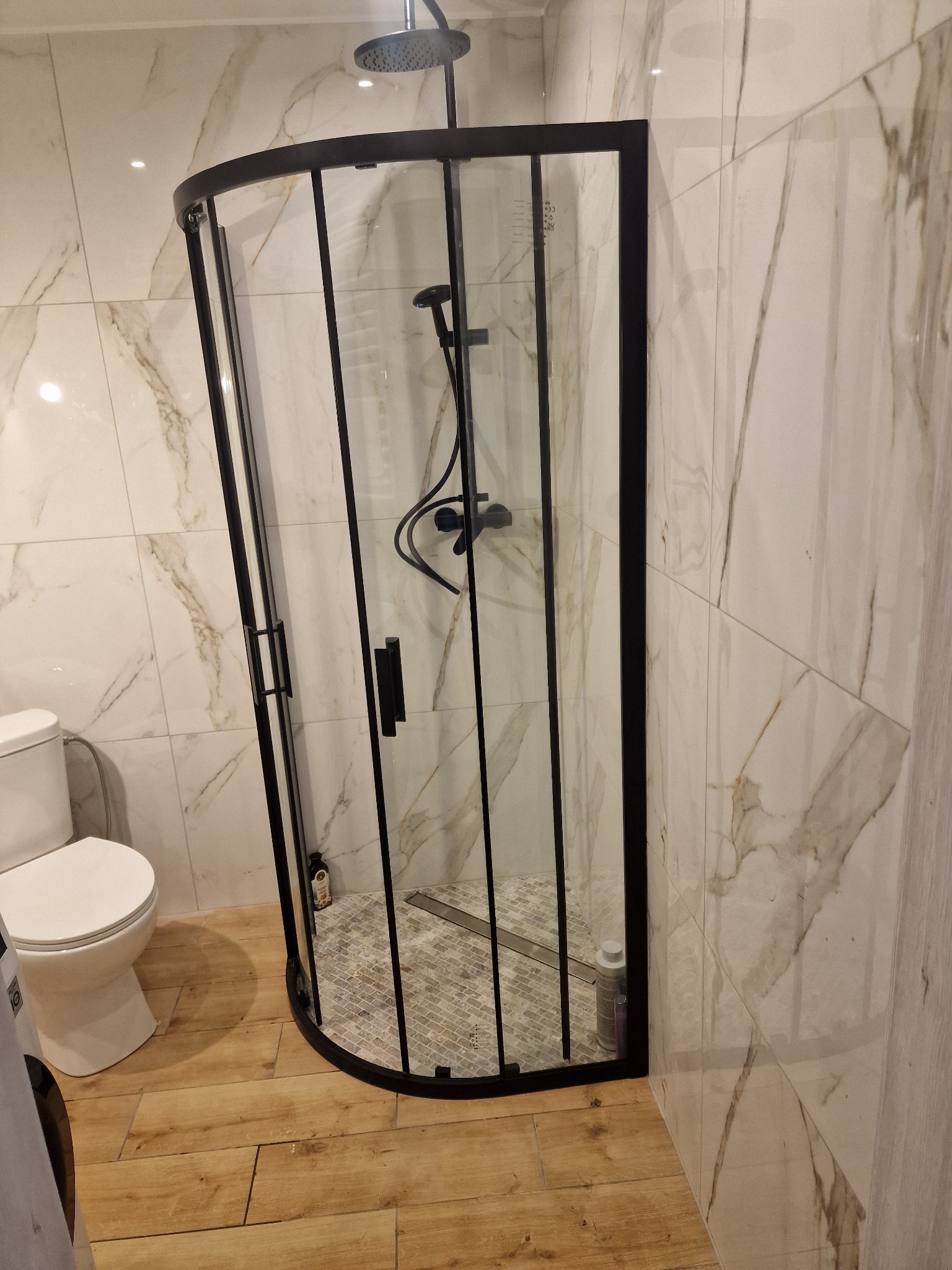 Nowoczesna kabina prysznicowa z czarnymi profilami, marmurowe płytki ścienne i drewnopodobna podłoga w łazience.